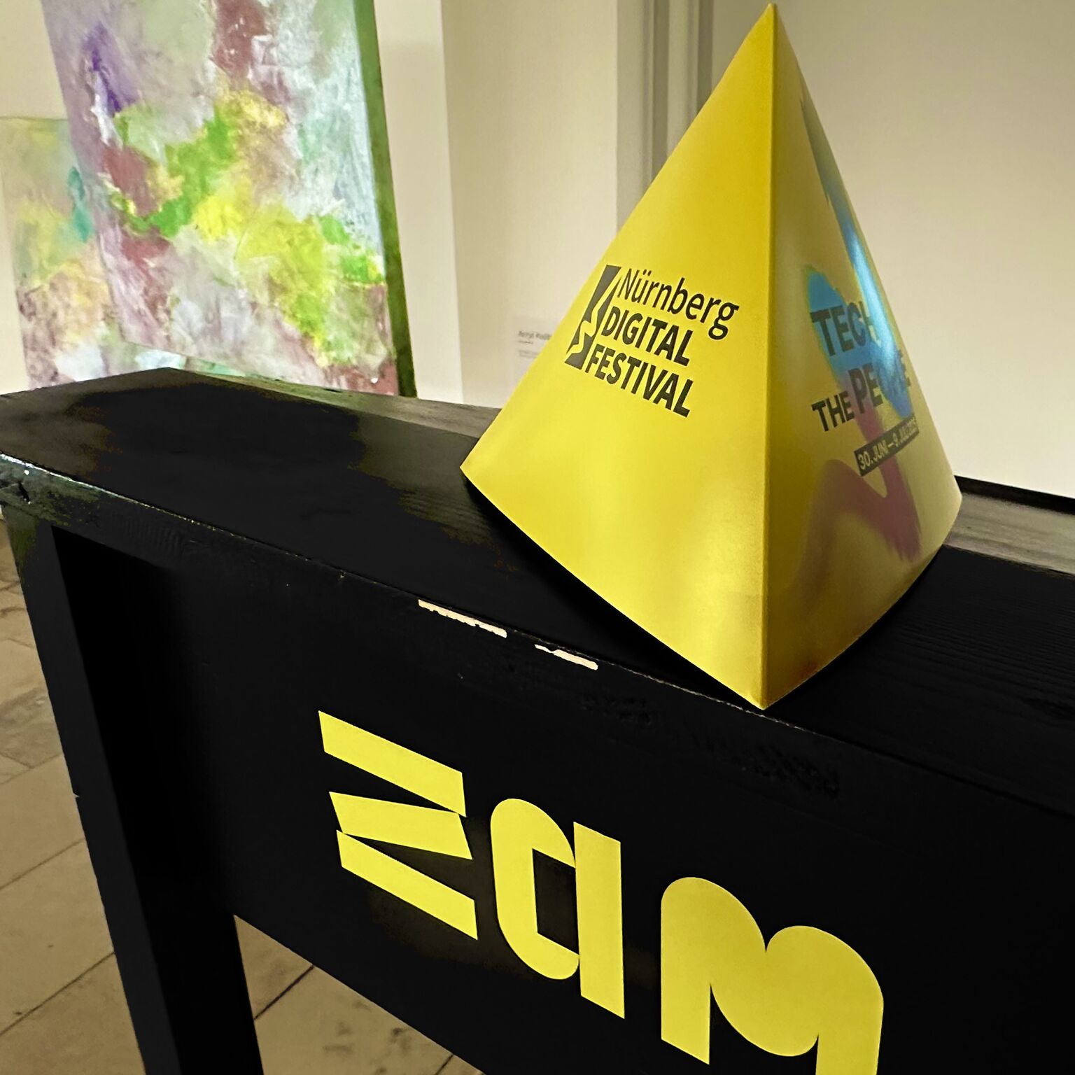 Im unteren Teil der Oberteil eines Stehpultes, schwarz lackiert, auf der Stirnseite ein Schriftlogo "ZAM" in gelber Farbe, das M leicht abgeschnitten. Auf dem Pult steht ein gelber dreiflächig-pyramidenförmiger Tischaufsteller, auf der linken sichtbaren Seite der Schriftzug Nürnberg Digital festival, auf der rechten Seite ein Logo mit geschwungenen Linien in den Farben blau und rot. Links im Hintergrund ist leicht verschwommen ein buntes Kunstwerk zu sehen, ansosnten ist weiße Wand sichtbar.