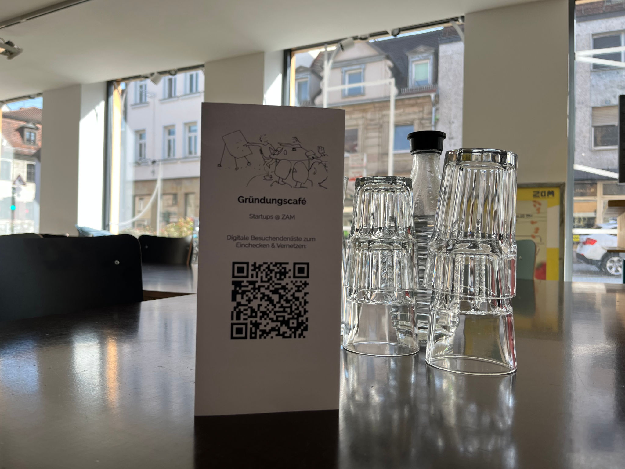 Im Vordergrund direkt die Fläche eines dunkelbraunen Tischs mit Tischaufsteller und der Beschriftung "Gründungscafé @ZAM" sowie einem QR-Code, daneben Wasserkaraffe und Gläser. Im Hintegrund Fenster, durch die die Gebäude