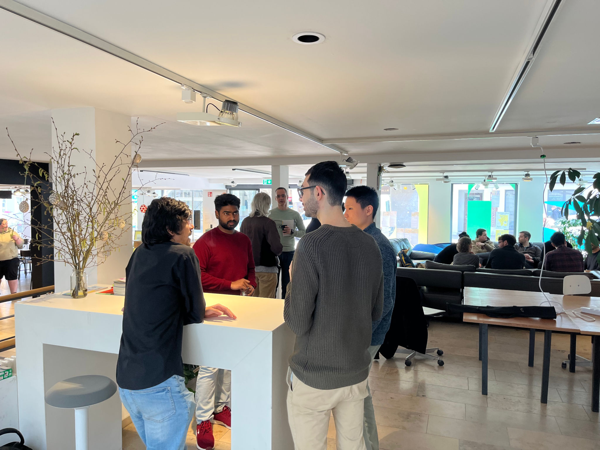 Im Vordergrund eine Gruppe von vier Menschen unterschiedlicher Herkunft an einem weißen Stehtpult, im Hintergrund Sofas auf denen sich weitere Menschen unterhalten. Ganz im Hintergrund große Fenster, aus denen man die Straße und die Gegenüberleigende Bebauung erkennt.