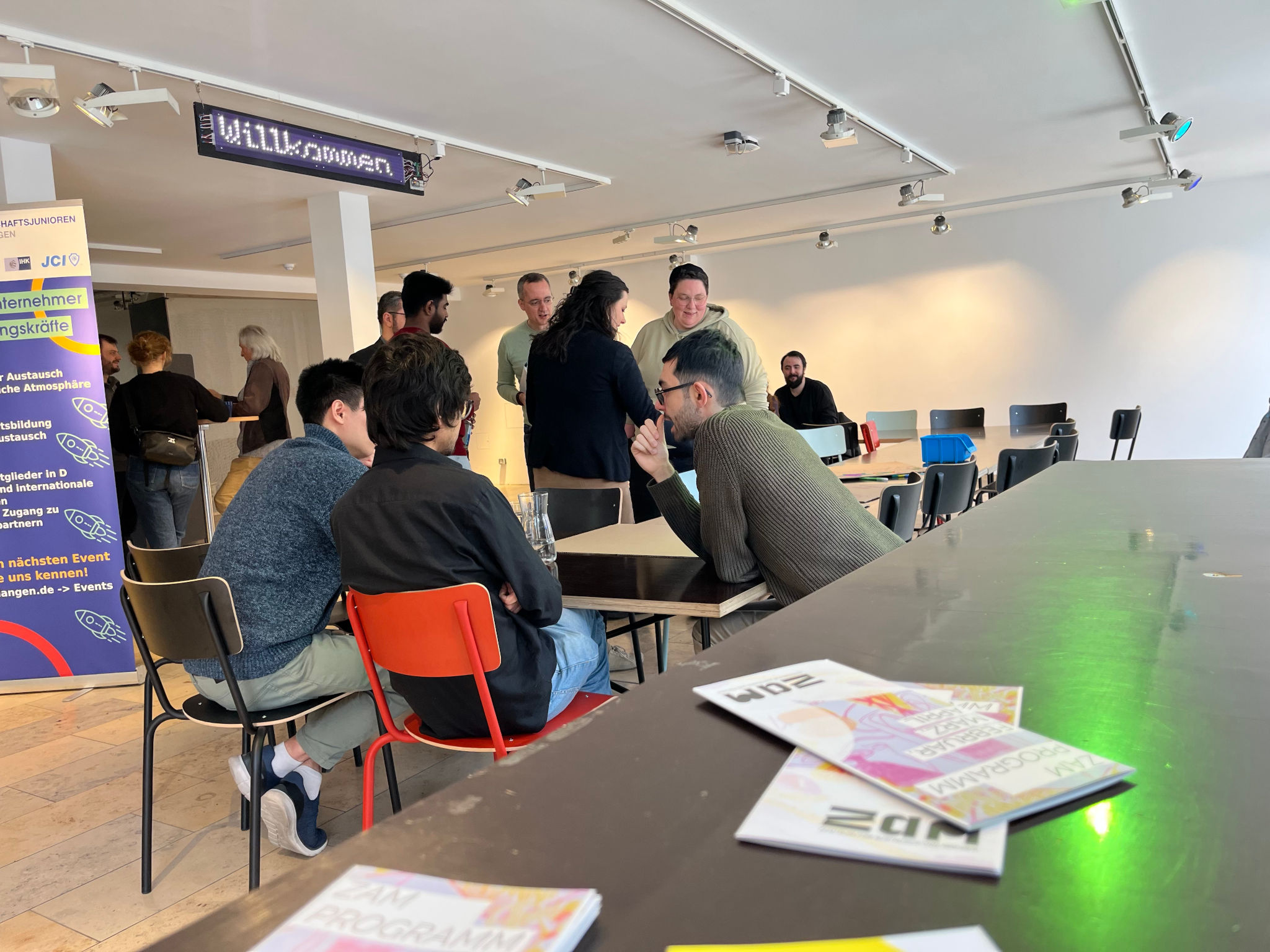 In der Mitte des Bildes eine Gruppe von Personen, die sich intensiv unterhält und auf unterschiedlichfarbigen, einfach aus Holzplatten und einem farbigen Metallgestenge gebauten, Stühlen an einem dunkelbraunen Tisch sitzt. Die Rechte untere Ecke zeigt den Fordergrund, ebenfalls eine Tischplatte auf denen Flyer liegen, auf diesen deutlich erkennbar das Logo des ZAM, stilisierte Buchstaben Z-A-M.