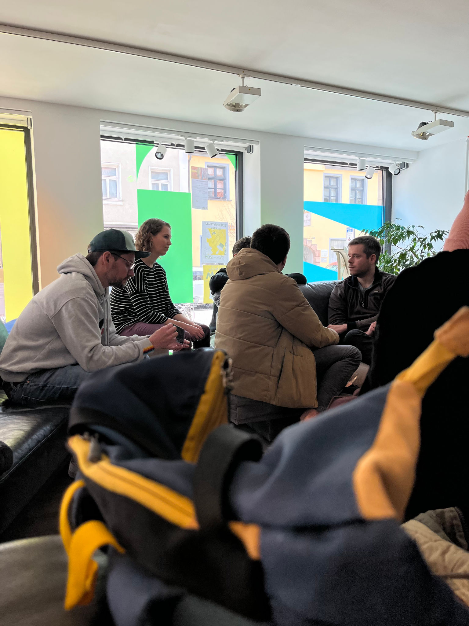 Gruppe von Menschen auf Sofas, die sich angeregt unterhalten. Im Hintegrund drei Fenster, auf denen jeweils ersichtlich etwas in bunter, halbdurchsichtiger Folie geklebt ist. Im unteren Bereich des Bildes sind farbige Teile eines Rucksacks erkennbar.