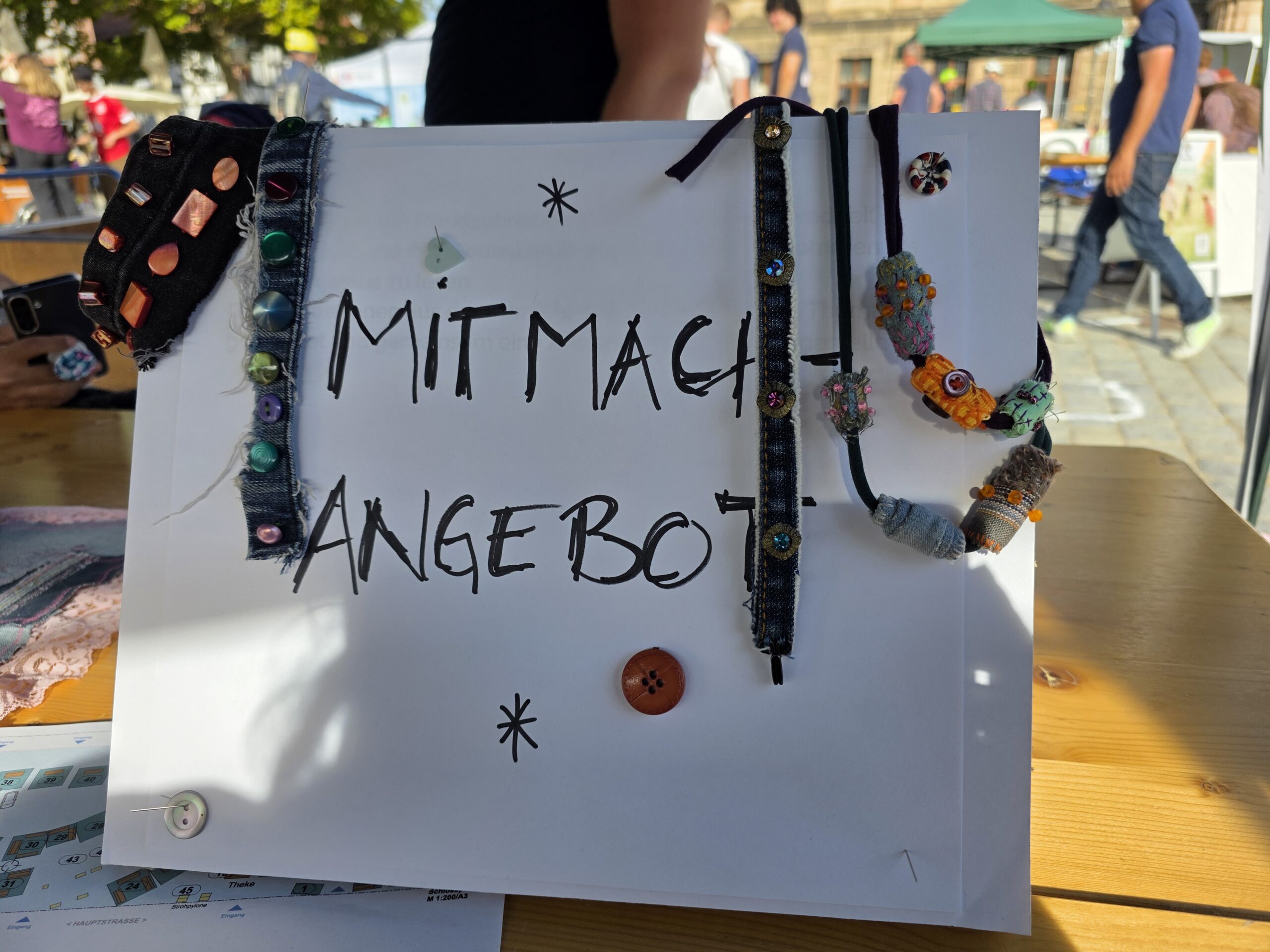 Ein Schild mit "Mitmach-Angebot", an dem verschiedene Stoffperlen hängen